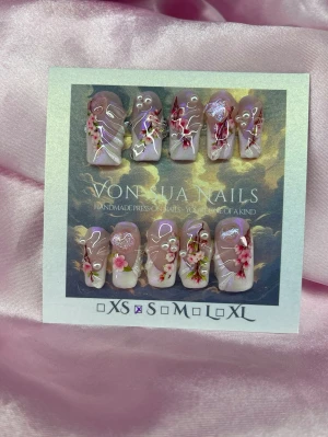 SakuraDrip (S);Medium Square - Handgjorda press-on naglar i storlek S: 15 mm, 11,5 mm, 12 mm, 11 mm, 9 mm . Naglarna har en nude bas med handmålad rosa french, sakura-blommor i rosa, vitt och mörkrosa, dekorerade med pärlor och skimrande detaljer, handmålad 3D nailart. Formen är medium lång square . Perfekt för dig som vill ha unika och kreativa naglar.