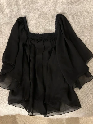 Svart luftig blus med vida ärmar - Snygg svart blus i luftigt material med vida ärmar och fyrkantig ringning. Den går att bära som off-shoulder eller normalt. Blusen har smockdetaljer upptill och knappar framtill vid halsen. Perfekt för en trendig och luftig look