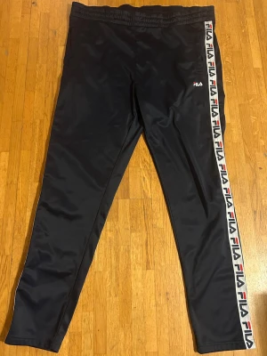 Marinblå byxor från FILA - Snygga marinblå byxor från FILA med logga längs hela sidan av benen. Byxorna har resår i midjan och är gjorda i ett glansigt, sportigt material. Perfekta för en avslappnad och trendig look.