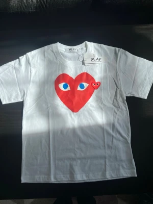 Vit Comme des Garçons Play T-shirt - Säljer en vit Comme des Garçons Play t-shirt med det ikoniska röda hjärtat med ögon på bröstet. Klassisk rund hals och korta ärmar. Tillverkad i mjuk bomull och har en clean, stilren look som passar perfekt till jeans eller shorts.