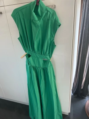 Grön långklänning med hög krage - Snygg grön långklänning med hög krage och markerad midja. Klänningen är ärmlös och har ett elegant, enkelt snitt som ger en stilren look. Perfekt för dig som vill sticka ut med färg. Materialet känns lätt och luftigt.