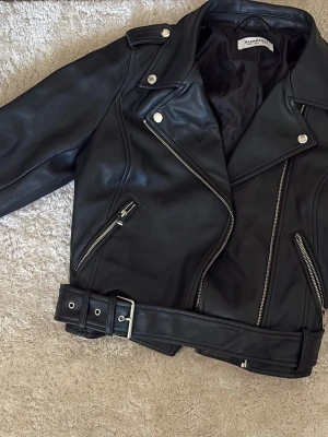 Svart skinnjacka från Nelly - Snygg svart skinnjacka från Nelly med klassisk bikerstil, silvriga dragkedjor och detaljer samt bälte i midjan. Jackan har bred krage och två snedställda fickor med dragkedja. Knappt använd 