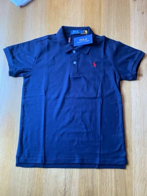 Marinblå piké från Polo Ralph Lauren - Klassisk marinblå pikétröja från Polo Ralph Lauren med röd broderad logga på bröstet. Skjortan har korta ärmar, krage och knappar framtill. Tillverkad i mjuk bomull som känns skön mot huden. Perfekt för en clean och stilren look.