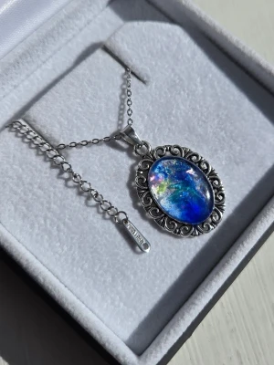 Halsband med blå sten och silverkedja - Unikt halsband med en oval, skimrande blå sten infattad i en detaljerad silverfärgad ram med snirkligt mönster. Kedjan är i rostfritt stål och har en justerbar längd. Efter att ha varit i vanligt dagsljus eller solljus så lyser hänget i ett starkt blått sken i mörker.  Perfekt för dig som gillar färgstarka accessoarer och vill sticka ut. Både kedjan och hänget tål vatten utan att missfärgas . 