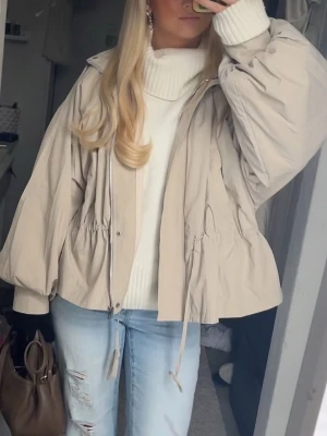 Beige jacka med snörning - Trendig och slutsåld beige jacka med dragkedja och snörning i midjan för en snygg siluett. Jackan har en hög krage och ballongärmar som ger en avslappnad och modern look. Perfekt för lager-på-lager och enkel att matcha med olika outfits. Perfekt nu till våren då den är tunn i materialet, funkar med både toppar, hoodies och stickade tröjor under.