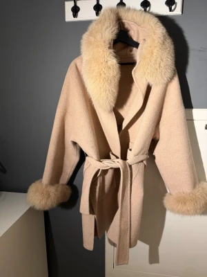 Beige kappa med fuskpälsdetaljer - Stilren beige kappa med stora fuskpälsdetaljer vid krage och ärmslut. Kappan har knytskärp i midjan och en avslappnad passform. Perfekt för dig som vill ha en trendig och mysig look under kalla dagar.
