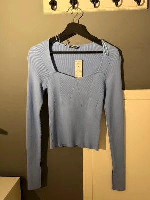 Ljusblå ribbad topp med lång ärm - Supersnygg ljusblå ribbad topp med lång ärm och fyrkantig ringning. Toppen har en figurnära passform och är tillverkad i ett stretchigt material som känns bekvämt mot huden. Perfekt att styla med jeans eller kjol för en trendig look.