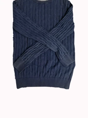 Mörkblå kabelstickad tröja från Gant - Mysig mörkblå kabelstickad tröja med rund halsringning och långa ärmar. Tröjan har ribbade muddar vid ärmslut och nederkant för extra komfort. Perfekt för kyliga dagar och enkel att matcha med jeans.