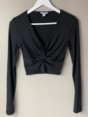 Svart croppad topp - Svart croppad topp med twistad detalj framtill. Toppen har lång ärm, ribbad struktur och djup v-ringning.
