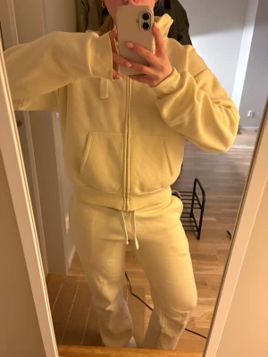 Gul mjukisdress med dragkedja - Säljer en ljusgul mjukisdress med hoodie och dragkedja framtill. Byxorna är i storlek s och kofta i m. Använd fåtal gånger