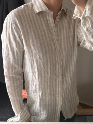 Beige och vit randig linneskjorta - Säljer en snygg beige och vit randig skjorta från John Henric . Skjortan är tillverkad i 100% linne, har klassisk krage, knäppning framtill och långa ärmar. Perfekt för en avslappnad men stilren look.