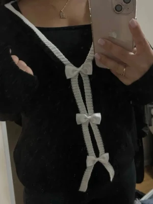 Söt tröja - Söt svart kofta med djup V-ringning och vita detaljer längs kanten. Tre vita rosetter framtill ger en gullig och trendig look. Perfekt att styla över ett linne eller t-shirt. Koftan har lång ärm och är stickad i mjukt material.