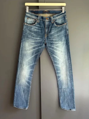 Blå slitna jeans från Nudie Jeans - Säljer ett par blå jeans från Nudie Jeans med snyggt slitna detaljer och klassisk femficksmodell. Jeansen har raka ben, orange sömmar och Nudie-logga på bakfickan. Tillverkade i denim med en cool tvättad look.