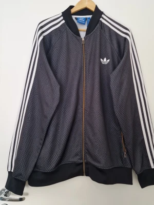 Svart tränjnhsjacka  från Adidas - Snygg svart grå träningsjacka från Adidas med klassiska vita ränder längs ärmarna och Adidas-logga på bröstet. Jackan har dragkedja framtill, ribbade muddar och ett diskret rutmönster i tyget. Perfekt för en sportig och avslappnad stil. Perfekt skick. Storlek 2XL