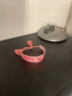Christian Dior rosa tygarmband - Säljer ett snyggt rosa och vitt tygarmband från Christian Dior med texten 'CHRISTIAN DIOR J'ADIOR' broderat i rött. Armbandet har justerbar knytning med tofsar i ändarna och är perfekt för dig som gillar exklusiva accessoarer med trendig vibe.