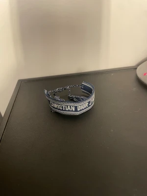 Blått Christian Dior tygarmband - Snyggt blått tygarmband från Christian Dior med texten 'J'ADIOR' och 'CHRISTIAN DIOR' i vitt. Armbandet har flätade band och tofsar i ändarna för justerbar passform. Perfekt accessoar för att lyfta din outfit med en exklusiv touch.