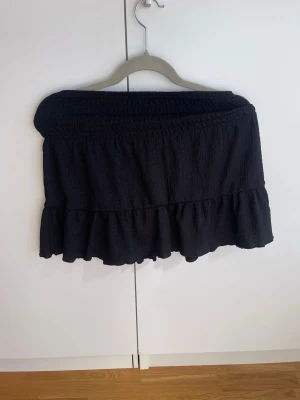 Svart volangkjol med resårmidja - Kort svart kjol med volangkant och bred resår i midjan. Kjolen har en lätt strukturerad yta och är perfekt för en trendig och avslappnad stil. Passar till många olika outfits och är enkel att matcha.