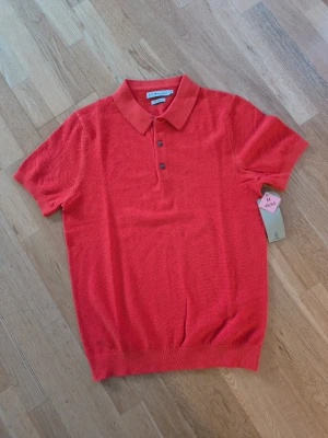 Orange pikétröja från S.C.W - Klassisk orange pikétröja från S.C.W med krage och tre knappar framtill. Tröjan har korta ärmar och ribbad nederkant. Tillverkad i stickad material som känns skönt mot huden. Perfekt för en avslappnad och stilren look.