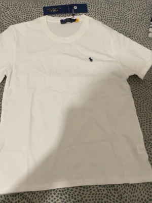 Vit t-shirt från Polo Ralph Lauren - Klassisk vit t-shirt från Polo Ralph Lauren med rund halsringning och den ikoniska broderade loggan på bröstet. Tillverkad i mjuk bomull för en skön och stilren look. Perfekt basplagg för alla tillfällen.