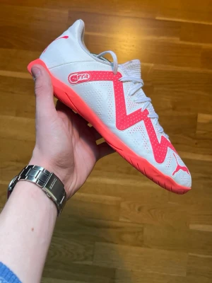 Vita och rosa Puma fotbollsskor - Säljer ett par snygga vita fotbollsskor från Puma med skarpa rosa detaljer och logga. Skorna har en låg profil, snörning och ovandel i syntetmaterial med perforeringar för bra ventilation. Sulan är också rosa och designad för inomhusspel.