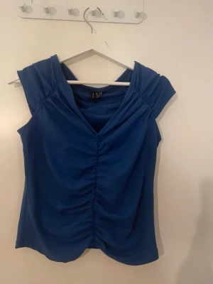 Blå v-ringad topp med rynk framtill - Snygg blå topp med v-ringning och rynkdetalj framtill. Ärmlös modell med mjukt, stretchigt material. Perfekt att styla med jeans eller kjol för en trendig look.