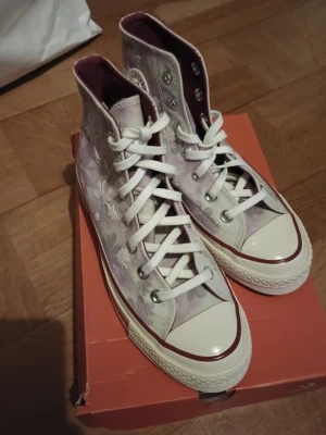 Converse All Star blommiga high tops Storlek 36 - Säljer ett par Converse All Star high tops med blommigt mönster i lila och vitt. Klassisk vit gummisula med röd rand och vita skosnören. Ikonisk Converse-logga på sidan och metallhål för snörning. Perfekta för dig som vill sticka ut med coola detaljer.