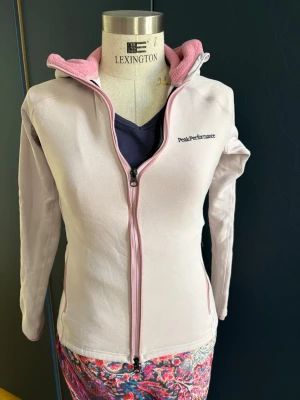 Ljusrosa hoodie Peak Performance - Rosa hoodie från Peak Performance. Insidan av huvan är fodrad i rosa fleece. Figurnära passform. Perfekt för chill dagar, träning eller som lager 2 under skidjackan.