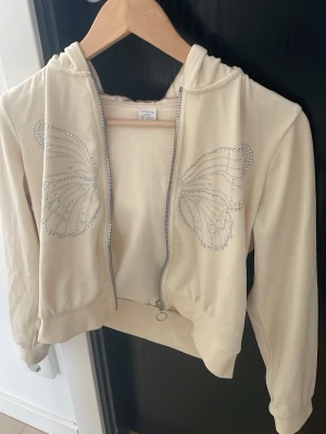 Beige hoodie med fjärilsdetaljer Lindex - Snygg beige hoodie från Lindex med dragkedja och huva. På bröstet finns två stora fjärilsvingar i silvriga strasstenar som ger en unik och trendig look. Perfekt för dig som vill sticka ut lite extra. Mjuk och skön bomullsblandning.