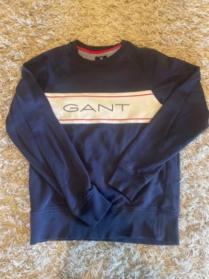 Marinblå sweatshirt från GANT - Snygg marinblå sweatshirt från GANT med vit panel och röda detaljer över bröstet. Stor GANT-logga framtill. Tröjan har rund halsringning och ribbade muddar vid ärmslut och nederkant. Perfekt för en avslappnad och stilren look.