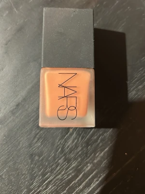 NARS flytande basprodukt persika - Flytande basprodukt från NARS i en frostad glasflaska med svart fyrkantigt lock. Färgen är varm persika och flaskan har tydlig logga på framsidan. Enkel att applicera och ger ett naturligt resultat.