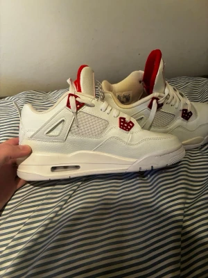 Vita Air Jordan 4 med röda detaljer - Säljer ett par vita Air Jordan 4 sneakers med röda detaljer och meshpaneler på sidorna. Skorna har vita skosnören, röd Jumpman-logga på plösen och röda plastdetaljer vid snörningen. Ovandelen är i läder och mesh, sulan är vit med synlig luftbubbla.