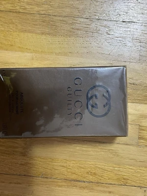 Gucci Guilty Absolute Pour Homme parfym - Gucci Guilty Absolute Pour Homme parfym i en lyxig brun förpackning med elegant logga och text i svart. Förpackningen är rektangulär och har en exklusiv känsla, perfekt för dig som gillar stilrena och klassiska dofter.