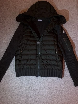 Moncler Tricot Cardigan  - Snygg svart dunväst från Moncler med stickade ärmar och ribbade muddar. Västen har en vadderad kropp, dragkedja framtill, två fickor med tryckknappar och en huva. Moncler-logga på ärmen och extra ficka med dragkedja på armen. Perfekt för kyliga dagar. Khaki Green, Kvitto medföljer 