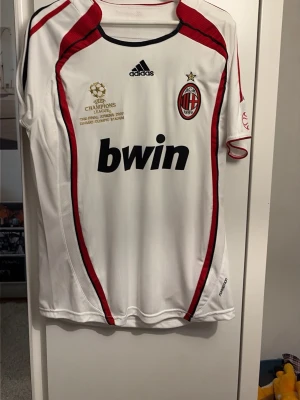 AC Milan Kaka' 22 Champions League tröja - Säljer en vit AC Milan matchtröja från Adidas med röda och svarta detaljer, Champions League-märke och Bwin-logga på bröstet. På ryggen står det KAKA' och nummer 22 i rött. Perfekt för fotbollsfans och samlare!