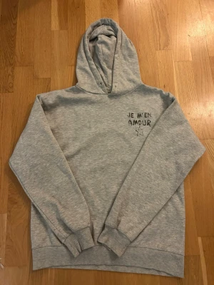 Grå hoodie med tryck 'JE M'EN AMOUR' - Säljer en grå hoodie med svart text 'JE M'EN AMOUR' och en stjärna tryckt både fram och bak. Hoodien har huva, ribbade muddar och är gjord i mjukt sweatshirtmaterial. Perfekt för en chill och avslappnad stil.