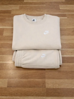 Beige Nike set med sweatshirt & byxor - Säljer ett beige set från Nike med matchande sweatshirt och byxor. Sweatshirten har rund halsringning och ribbade muddar, båda plaggen har Nike-logga broderad i vitt. Mjukt material, perfekt för en avslappnad stil.