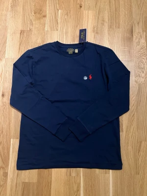 Ralph Lauren Tröja - Snygg och klassisk mörkblå långärmad tröja från Ralph Lauren med den ikoniska röda loggan på bröstet. Perfekt för både vardag och uppklädd stil. PRIS KAN DISKUTERAS!