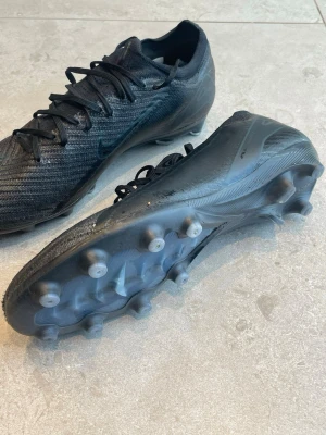Svarta Nike Mercurial Vapor 16 fotbollsskor - Säljer ett par svarta Nike Mercurial fotbollsskor med snörning och mönstrad ovandel i syntetmaterial som kallas gripknit. Skorna har fasta dobbar för spel på konstgräs och en slimmad, modern design. Kommer med en svart Nike-påse med tryck. Perfekta för dig som vill ha grym touch och speed på planen.