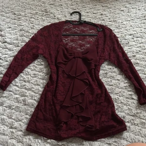 Vinröd spetsblus med stor rosett - Vinröd blus i spets med lång ärm och stor dekorativ rosett framtill. Blusen har djup urringning och volangdetaljer längs rosetten. Materialet är genombruten spets som ger en elegant look. Perfekt för dig som vill sticka ut med en unik detalj.