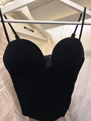 Svart korsettopp med smala band - Snygg svart korsettopp med smala axelband och markerade kupor. Toppen har en dragkedja framtill och en figurnära passform som framhäver siluetten. Perfekt att styla med höga jeans eller kjol för en trendig look.