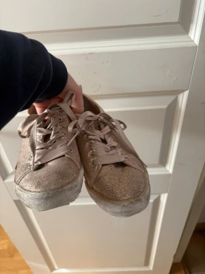 Glittriga sneakers från Guess - Snygga sneakers från Guess i glittrigt roséguld med vita sulor och matchande skosnören. Skorna har låg modell och är perfekta för dig som vill sticka ut med lite extra bling. Märkeslogga bak på hälen och glittrigt material över hela skon.