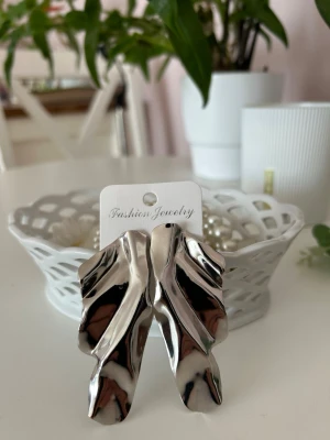Silvriga vågiga örhängen - Stora, statement örhängen i blankt silverfärgat metallmaterial med vågig, abstrakt form. Ger en cool och modern look och passar perfekt för dig som vill sticka ut med accessoarerna. Märkta med Fashion Jewelry.