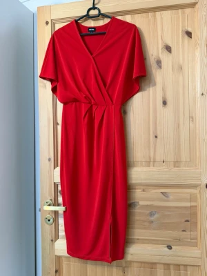 Röd omlottklänning med slits - Snygg röd omlottklänning med v-ringning och korta ärmar. Klänningen har en markerad midja och en slits framtill som ger en trendig look.  Tillverkad i mjukt och följsamt material.