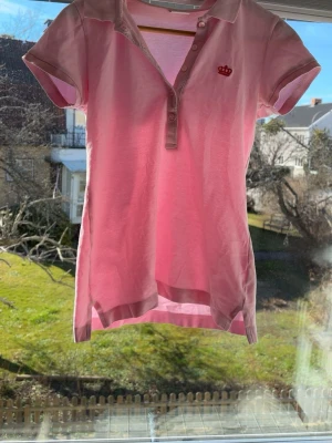 Rosa pikétopp från Gina Tricot/ storlek s fast passar xs!! - Säljer en ljusrosa pikétopp från Gina Tricot med korta ärmar och knappar framtill. Toppen har en liten broderad krona på bröstet och klassisk krage. Perfekt till jeans eller kjol för en avslappnad look.