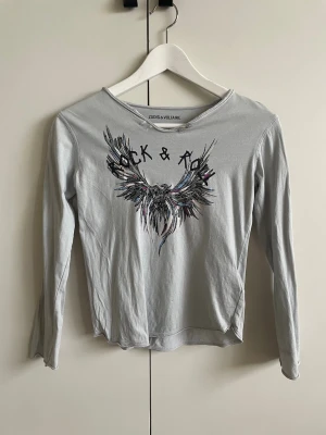 Grå långärmad topp Zadig & Voltaire - Snygg grå långärmad topp från Zadig & Voltaire med tryck av vingar och texten 'Rock & Roll' framtill. Rund halsringning med två knappar vid axeln ger en cool detalj. Perfekt för dig som gillar edgy och avslappnad stil.