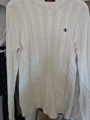 Offwhite kabelstickad tröja Morris - Kabelstickad offwhite tröja från Morris med rund halsringning och diskret blå logga broderad på bröstet. Tröjan har långa ärmar och ribbade muddar vid ärmslut och nederkant.Perfekt för en clean och klassisk look. Storlek XS. I Bomull passar perfekt till vår och sommar.