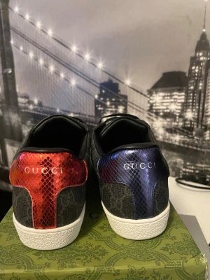 Gucci sneakers med GG-mönster - Snygga Gucci sneakers med klassiskt GG-mönster i grått tyg. Baksidan har metallicröda och metallicblå detaljer i ormskinnslook, båda med Gucci-logga. Skorna har svarta läderdetaljer och vita sulor. Perfekta för dig som vill sticka ut med exklusiva detaljer.