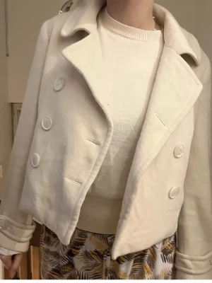 Vit kort jacka med stora knappar - Stilren vit jacka med kortare passform och stora dekorativa knappar framtill. Jackan har bred krage och långa ärmar, perfekt för att lyfta en outfit. Passar dig som gillar en klassisk men trendig look.