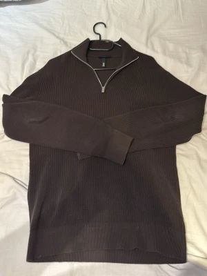 Brun ribbstickad tröja från H&M - Mörkbrun ribbstickad tröja från H&M med halv dragkedja och hög krage. Tröjan har långa ärmar och en stilren, enkel design som passar perfekt till lager på lager. Materialet ser ut att vara mjukt och bekvämt.