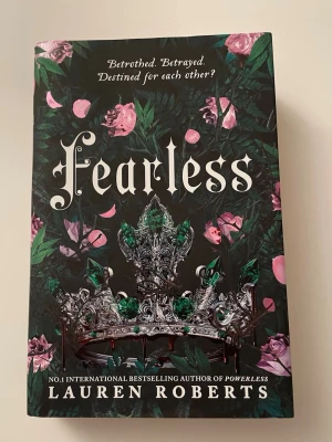 Fearless Collector’s Edition  - Säljer boken 'Fearless' av Lauren Roberts. Omslaget är mörkt med gröna blad, rosa rosor och en detaljerad krona i silver och grönt. Själva boken har ett ljusrosa hårdband med silvermönster av en krona och rosor. Perfekt för dig som gillar fantasy och romantik.